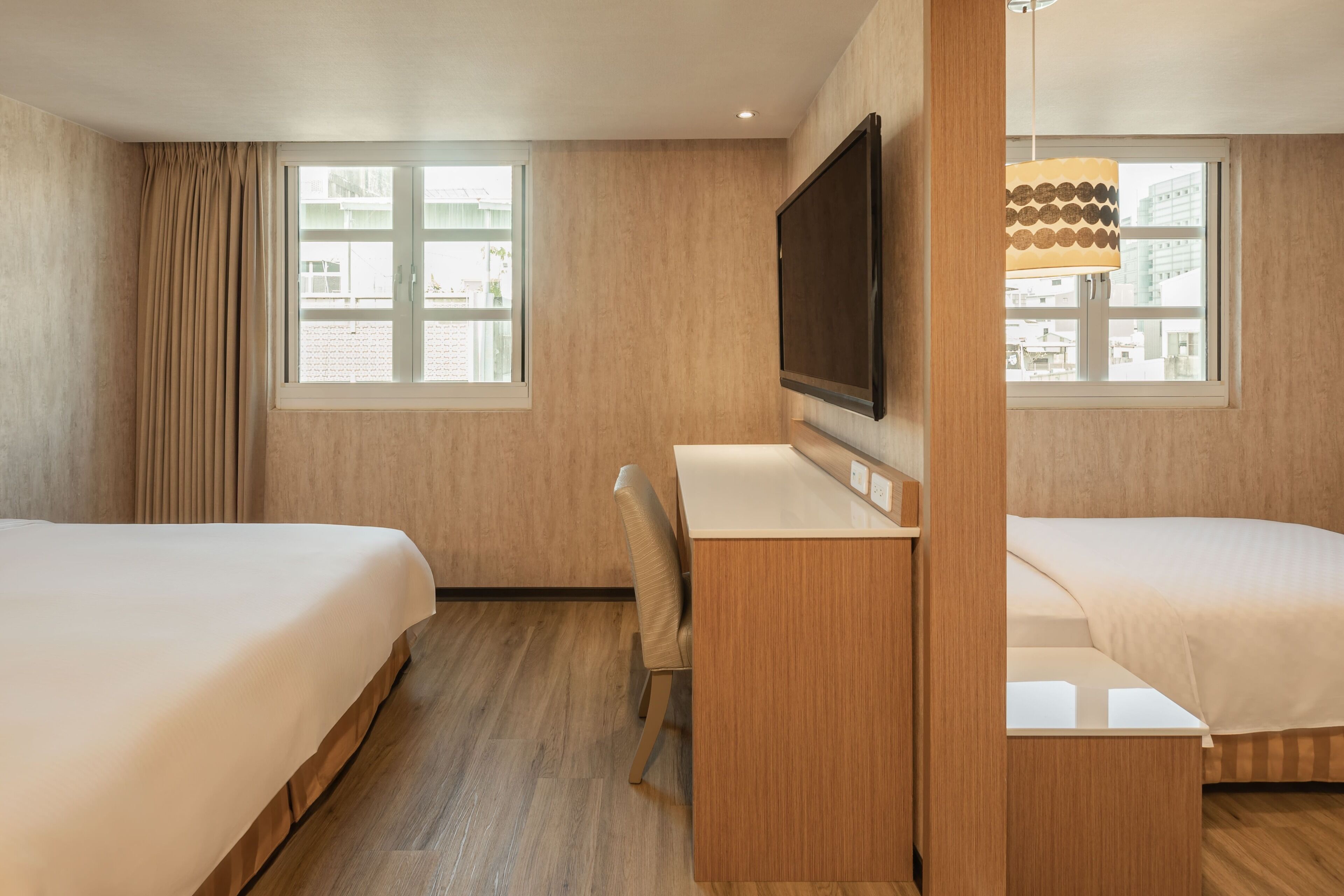 A hotel room with two beds, a desk, a chair, a TV, and large windows.