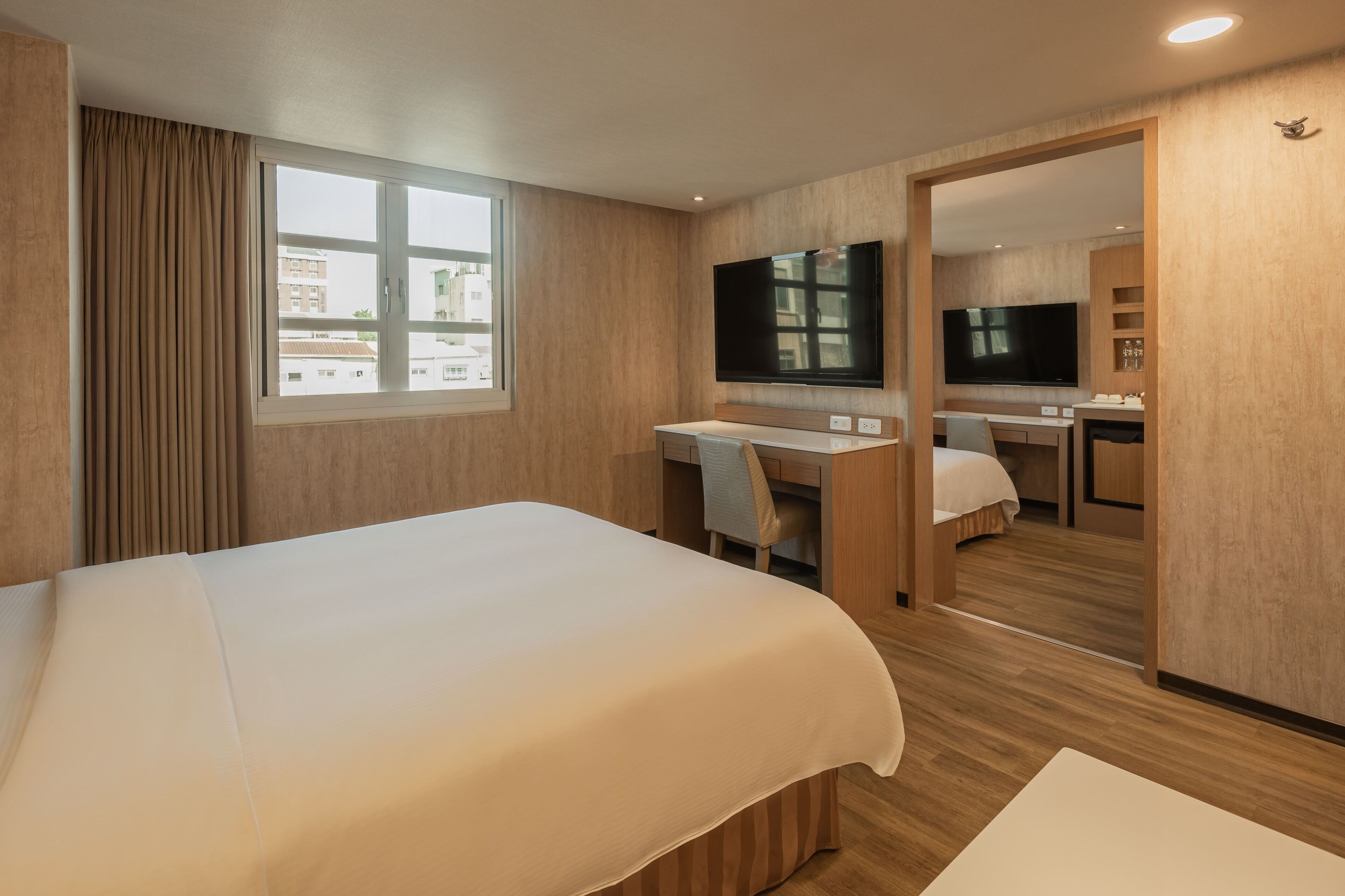A hotel room with a large bed, a desk, a chair, a TV, and a view of the city.
