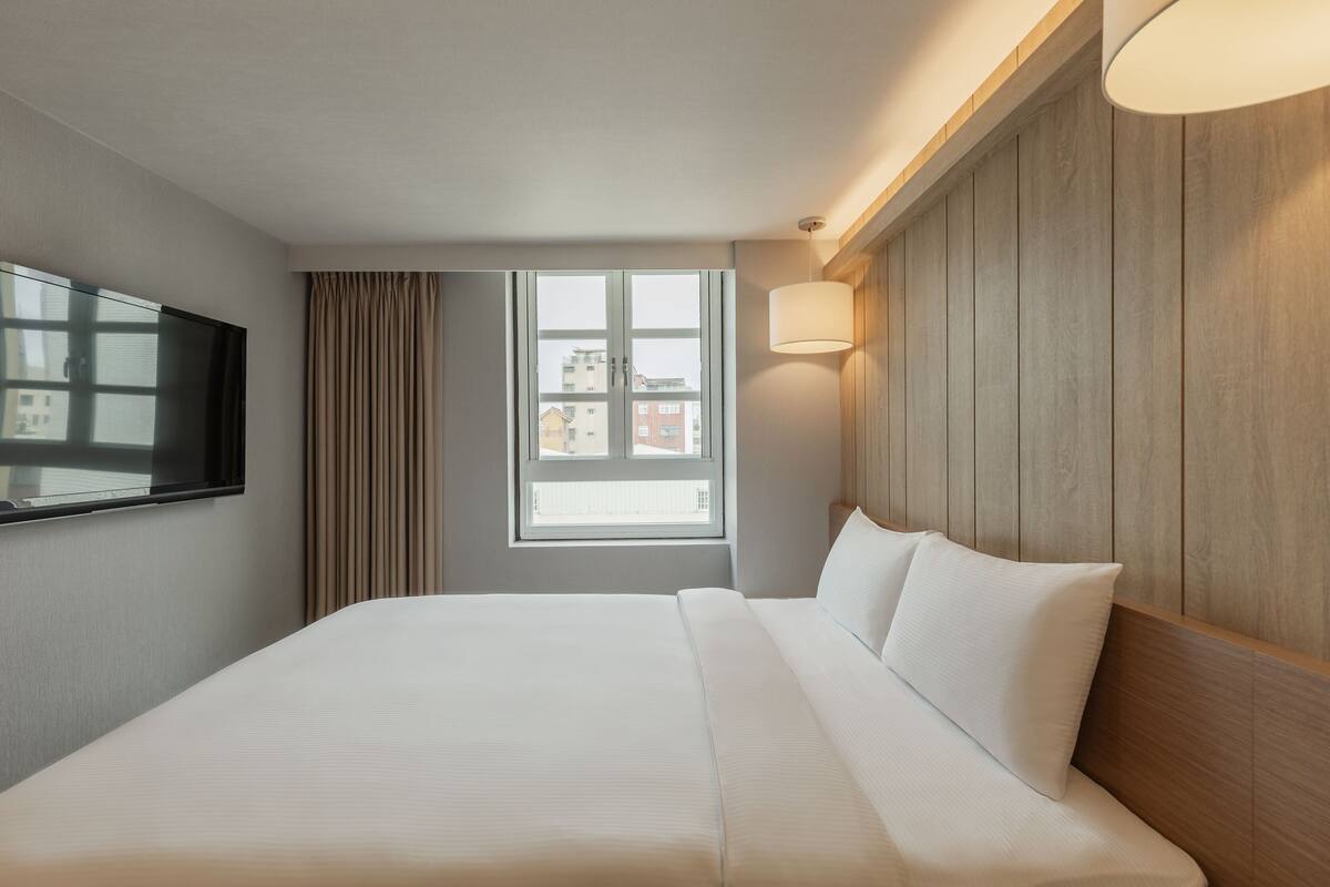 Ein Hotelzimmer mit einem großen Bett, einem flachbildfernseher an der Wand, einem Fenster mit Vorhängen und einem hölzernen Kopfteil.