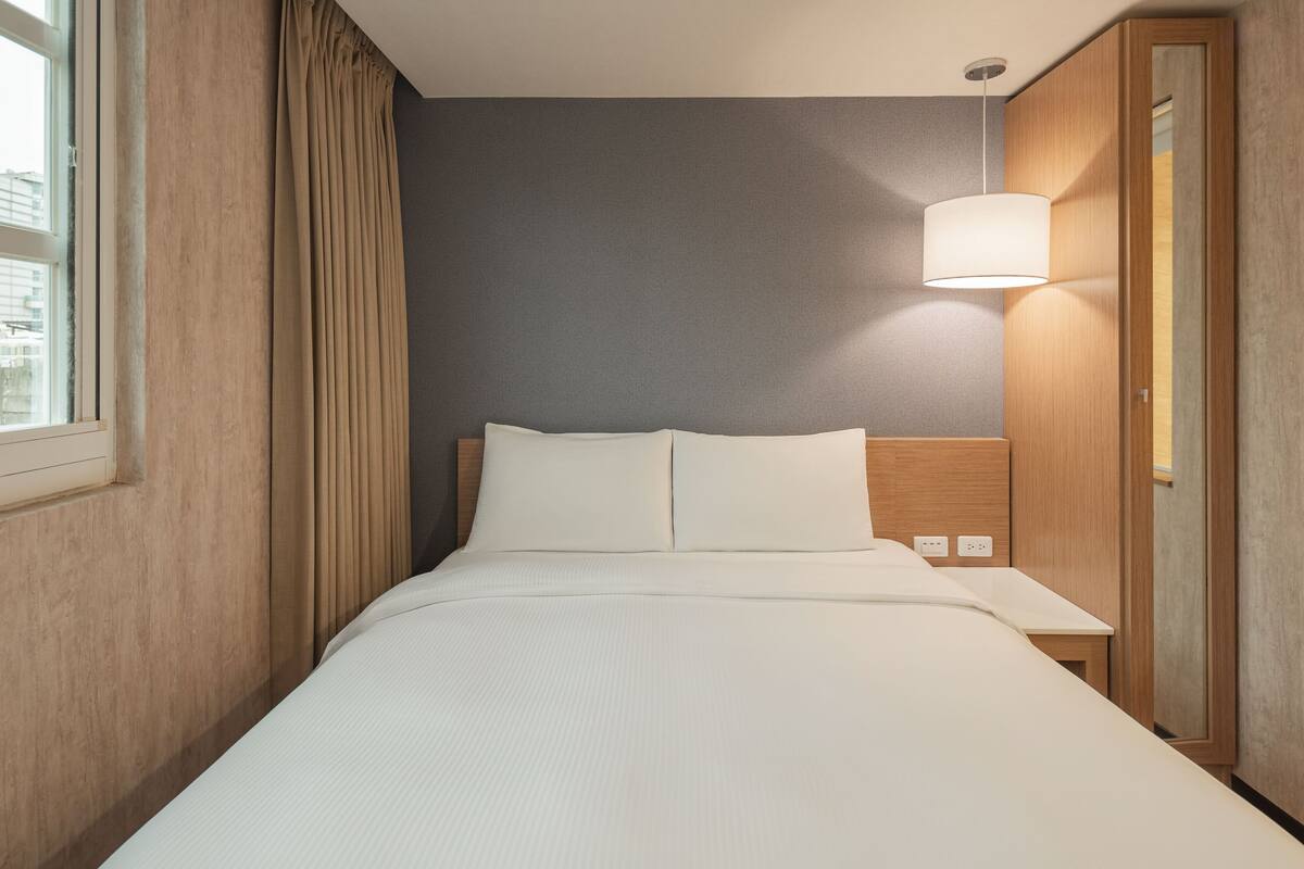 Ein Hotelzimmer mit Bett, Nachttisch, Lampe und einem Fenster mit Vorhängen.
