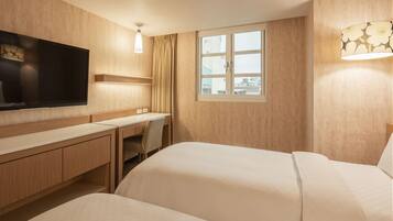 A hotel room with a large flat-screen TV, a desk, two beds, and a window with a view of buildings.