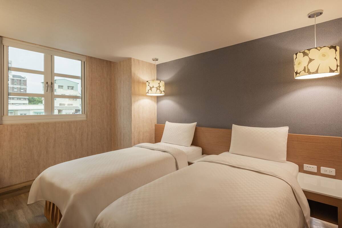 Ein Hotelzimmer mit zwei Betten, einem Fenster, einer Wandleuchte und einem hölzernen Kopfteil.
