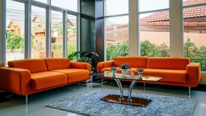A modern living room with two orange sofas, a glass coffee table, and large windows offering a view of a red-tiled roof and greenery. - Excella Hotel (Ubon Ratchathani)