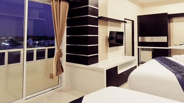 A modern hotel room with a large bed, a TV, a minibar, and a view of the city at night. - Excella Hotel (Ubon Ratchathani)