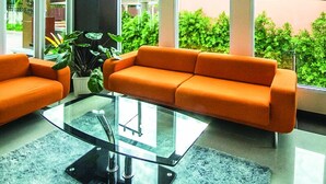A modern living room with a glass coffee table, orange sofa, and large windows offering a view of greenery and a red-roofed building. - Excella Hotel (Ubon Ratchathani)