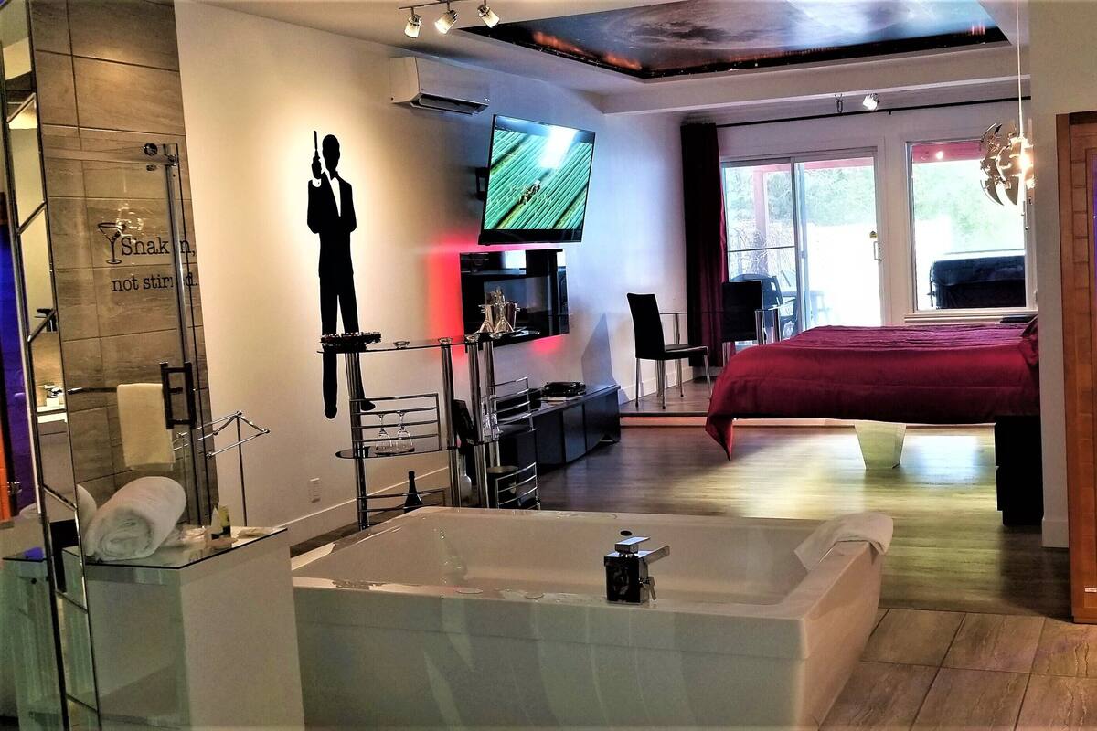 Una habitación de hotel moderna con una cama grande, un baño separado con bañera, un televisor montado en la pared y vistas al exterior a través de las ventanas.