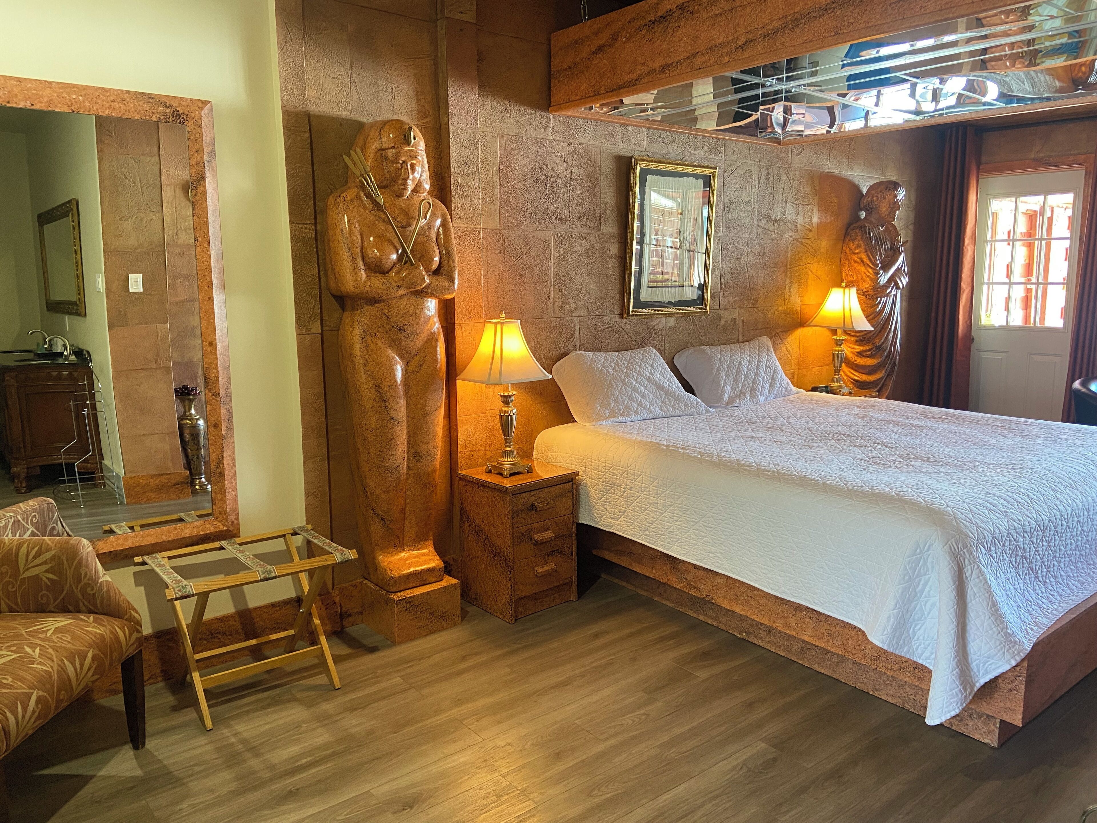 A spacious bedroom with a large bed, a nightstand with a lamp, a wooden chair, and a statue.