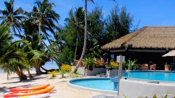 A pool area with a thatched-roof structure, palm trees, and kayaks.
