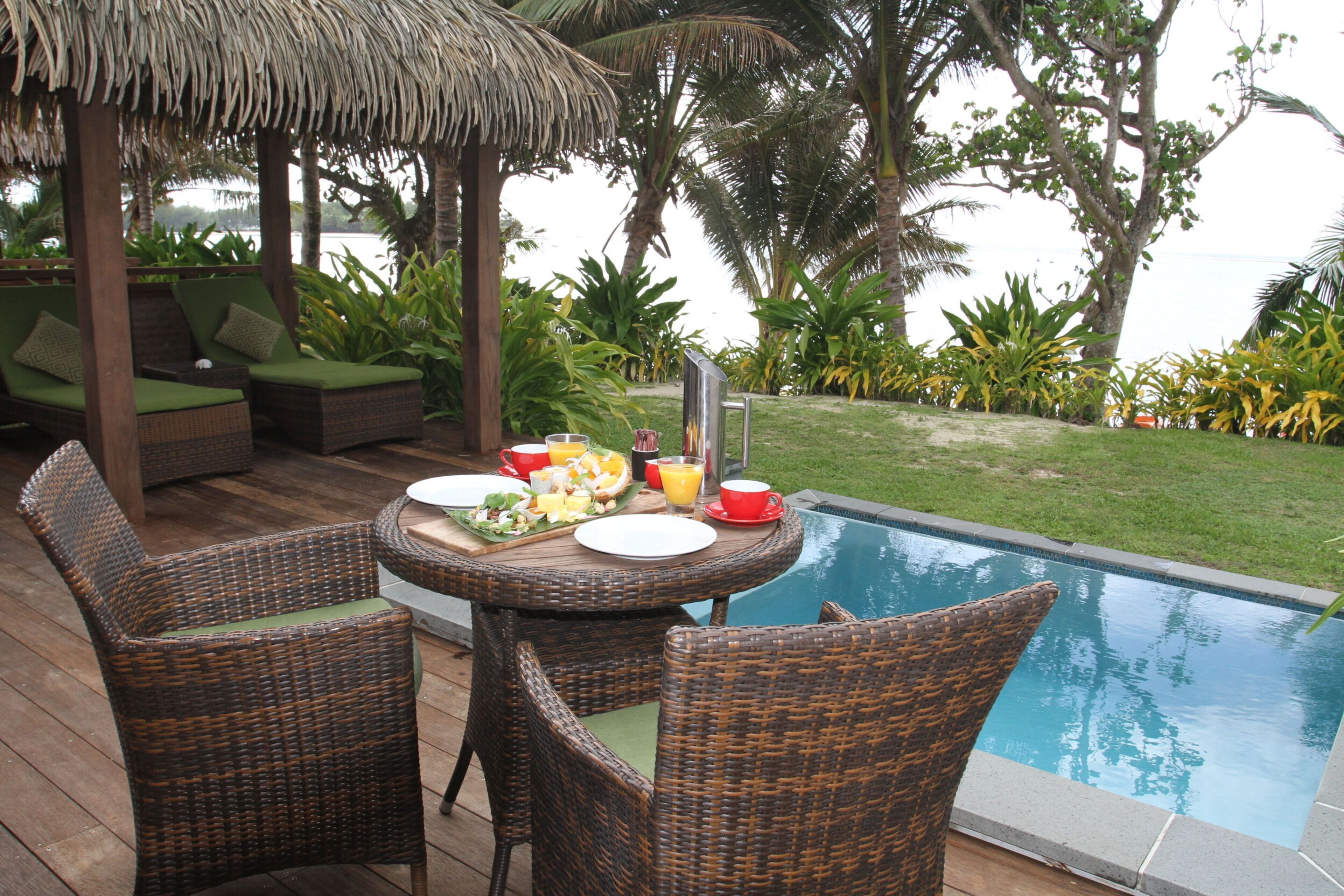 A poolside dining area with wicker furniture, a table set for breakfast, and a view of the pool and surrounding greenery.