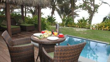 A poolside dining area with wicker furniture, a table set for breakfast, and a view of the pool and surrounding greenery.
