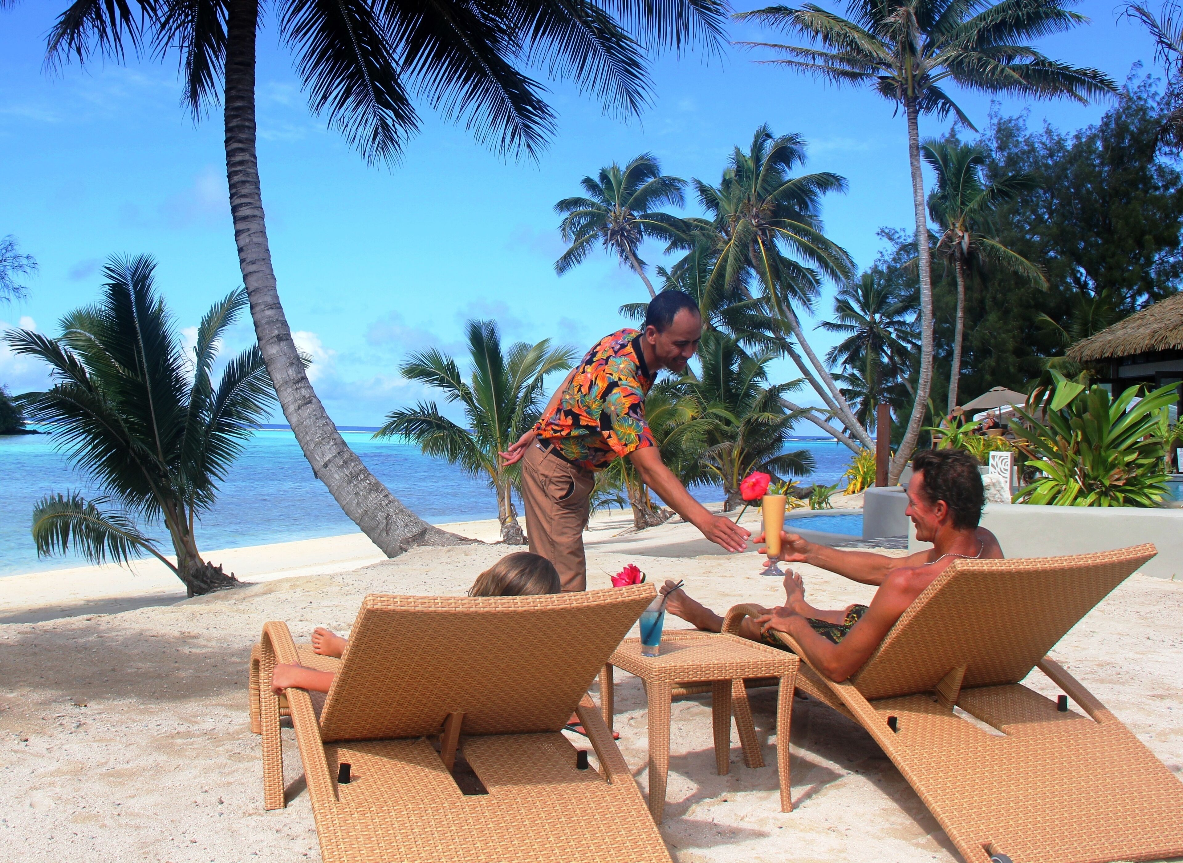 Un entorno en la playa con sillones de descanso y una mesa, palmeras y una vista despejada del océano.