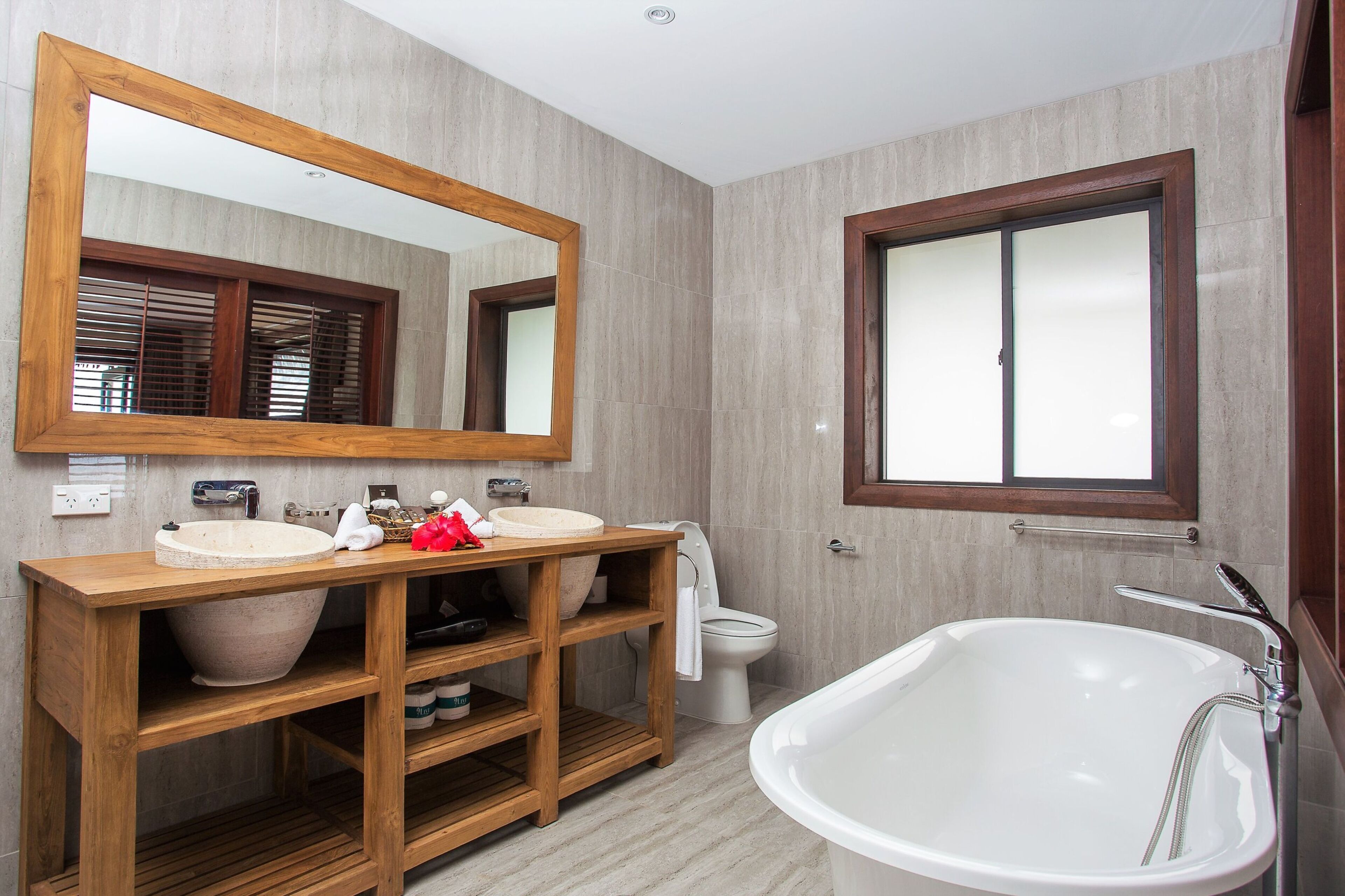 Un baño moderno con tocador de madera, dos lavamanos, un espejo grande, una bañera y una ventana con persianas.