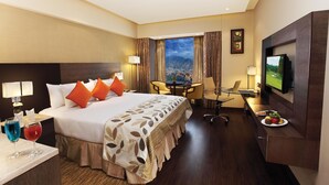A hotel room with a large bed, a desk with a computer, a TV, and a view of a cityscape through the window. - Hotel Vrisa (Jaipur)