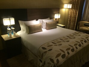 A hotel room with a large bed, two bedside tables, a nightstand, a lamp, a chair, and a window with curtains. - Hotel Vrisa (Jaipur)