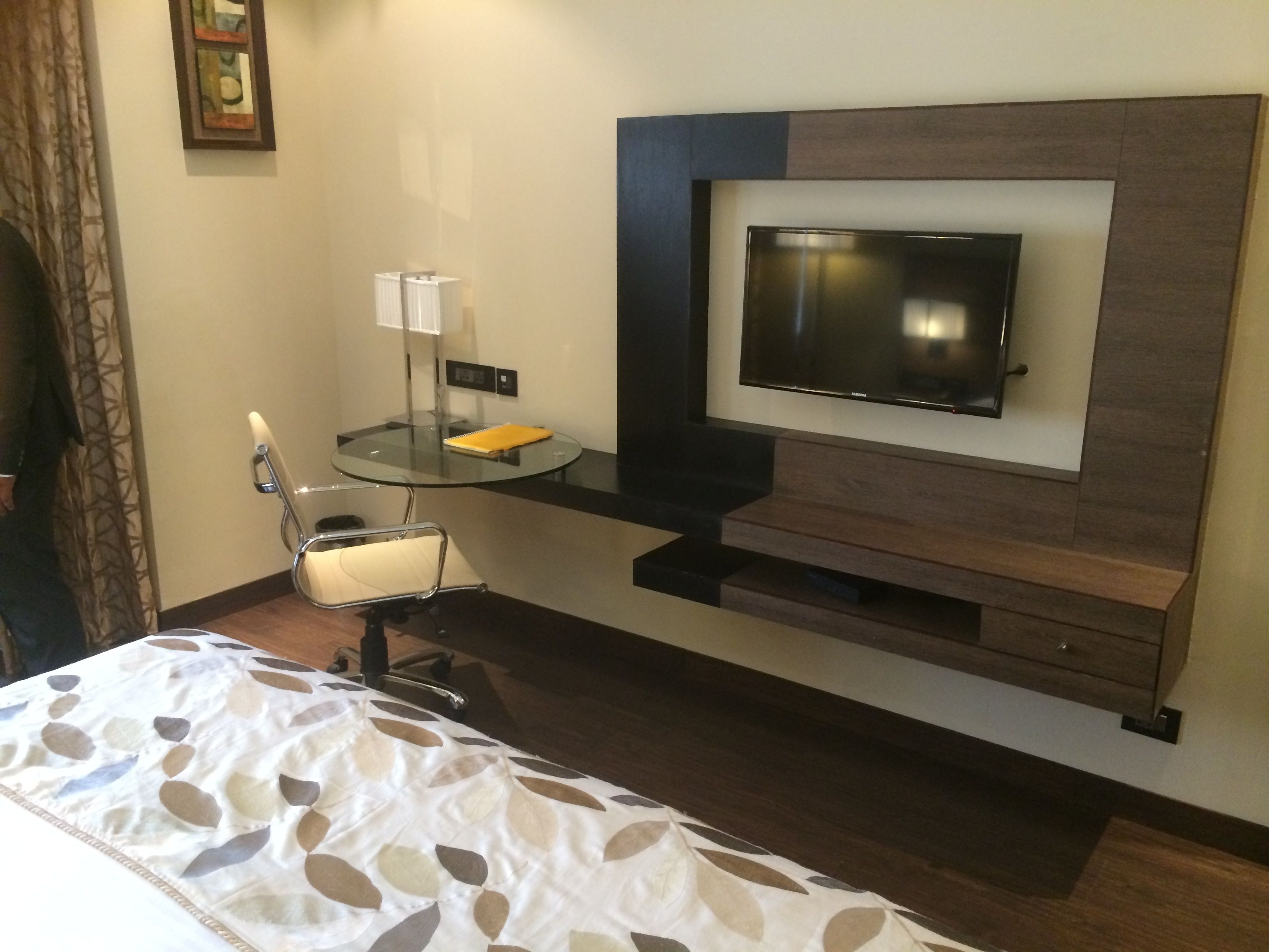 Habitación de hotel con cama, escritorio y silla, televisor de pantalla plana en la pared, y una mesita de noche con lámpara.