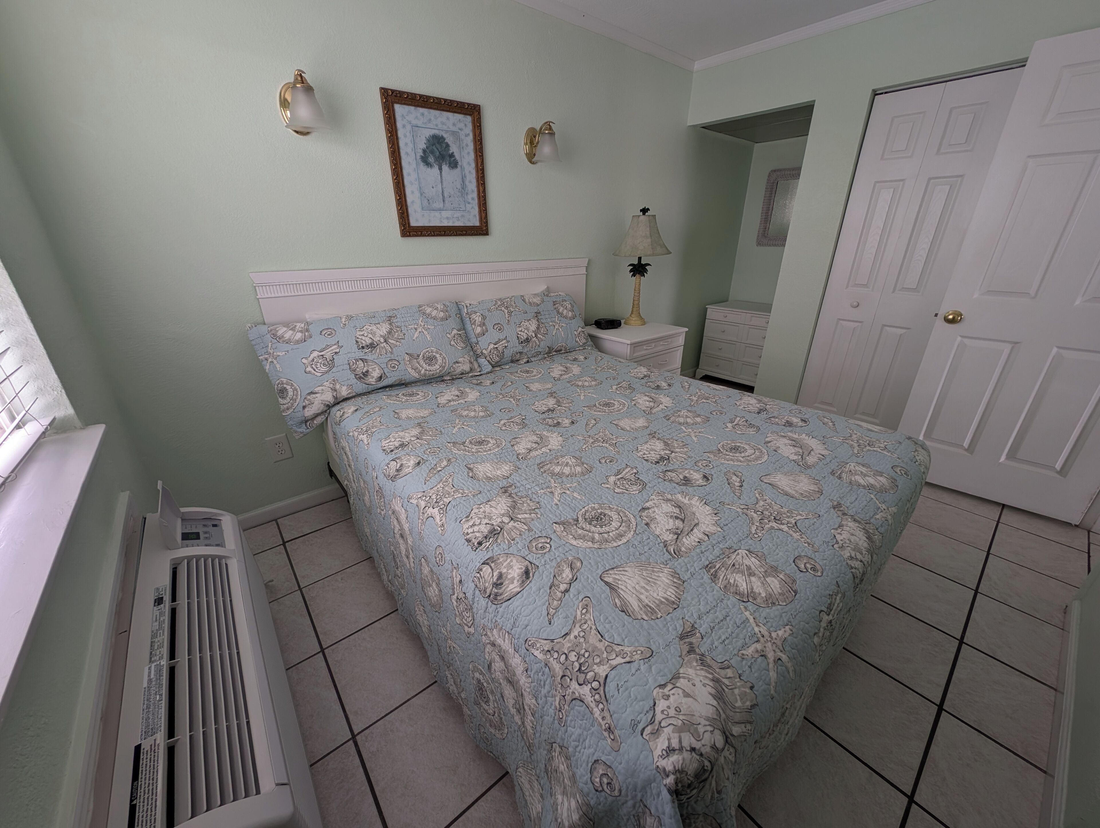 A bedroom with a bed, a nightstand, a lamp, a heater, a door, and a wall-mounted picture.
