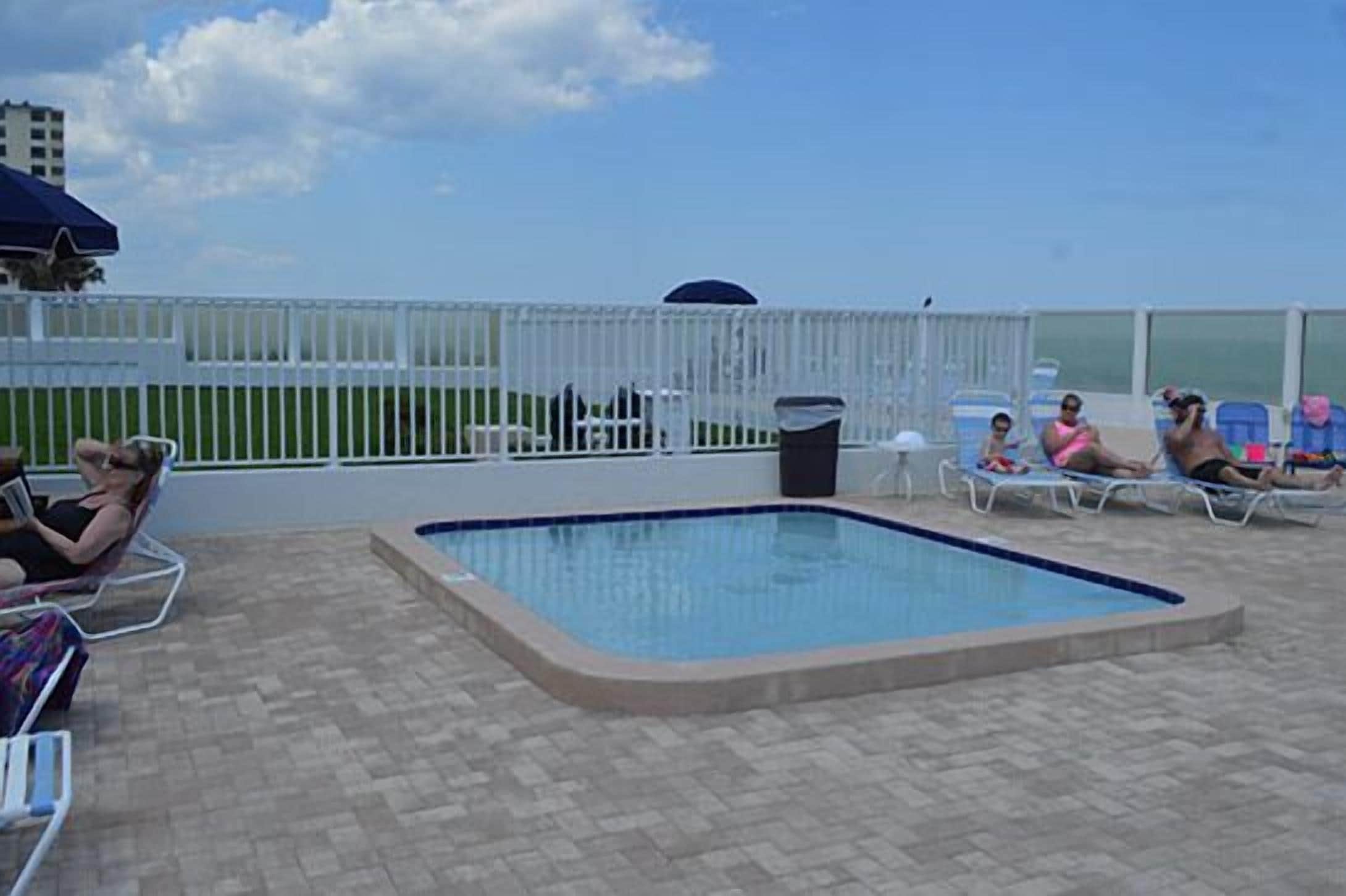 A rooftop pool area with a small pool, lounge chairs, and a white fence.