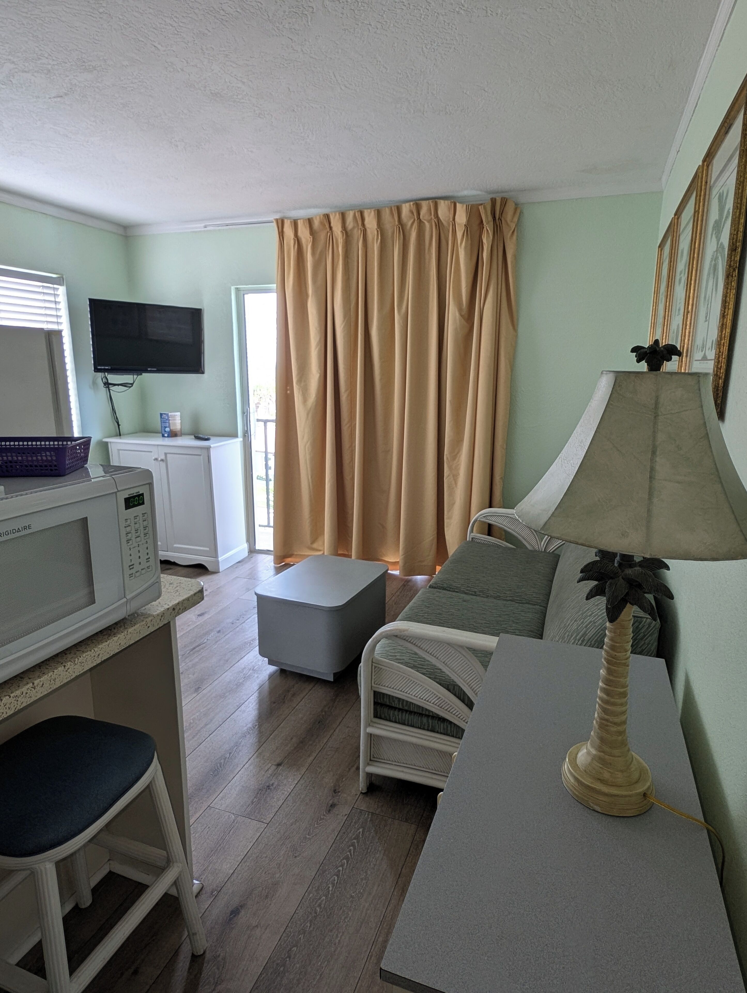 A compact living space with a kitchenette, a flat-screen TV, a sofa, a lamp, and a window with curtains.