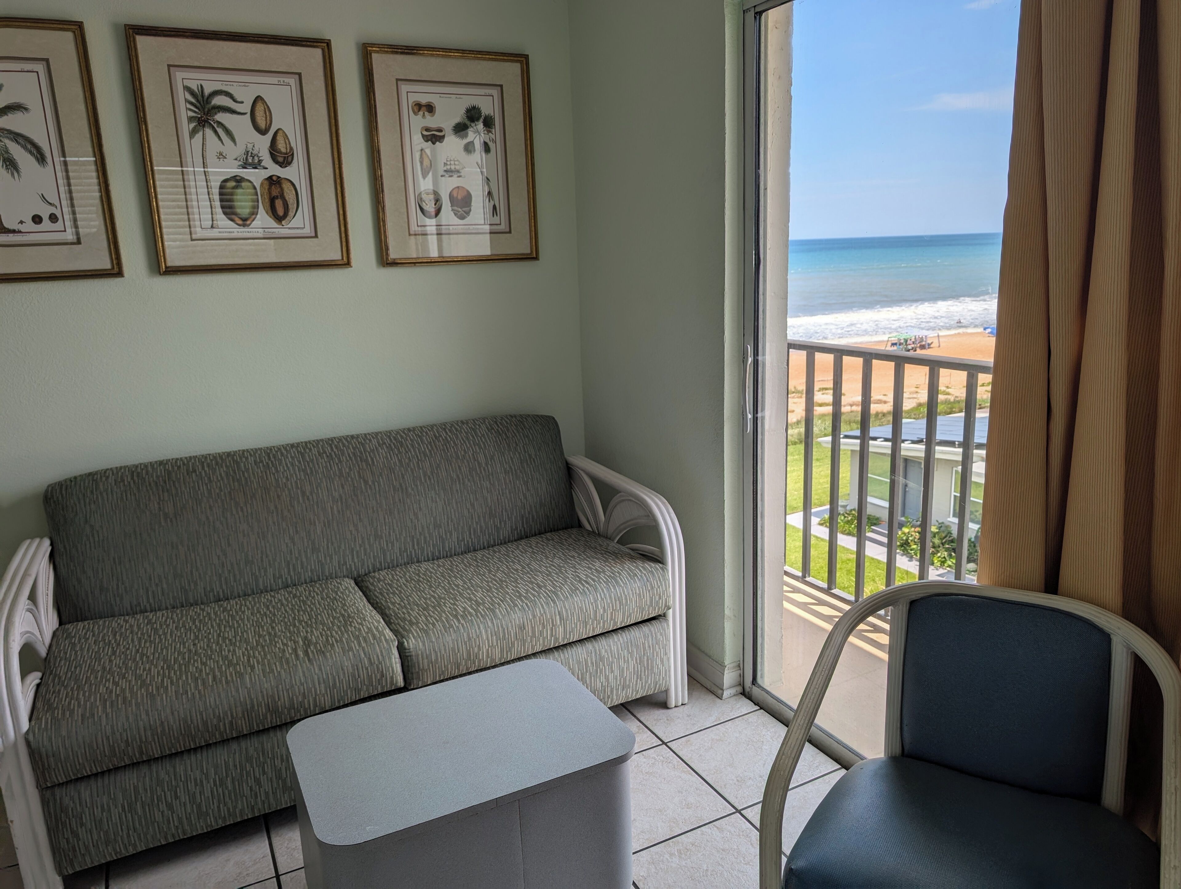 Un balcon donnant sur la plage, un canapé gris, une petite table et, au mur, des reproductions encadrées de motifs botaniques.