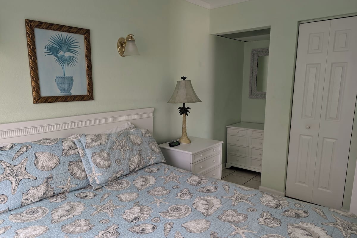 Un dormitorio con una cama que presenta un estampado de conchas marinas y estrellas de mar en azul y blanco, una lámpara de noche, un cuadro con una planta en maceta y un aparador blanco.