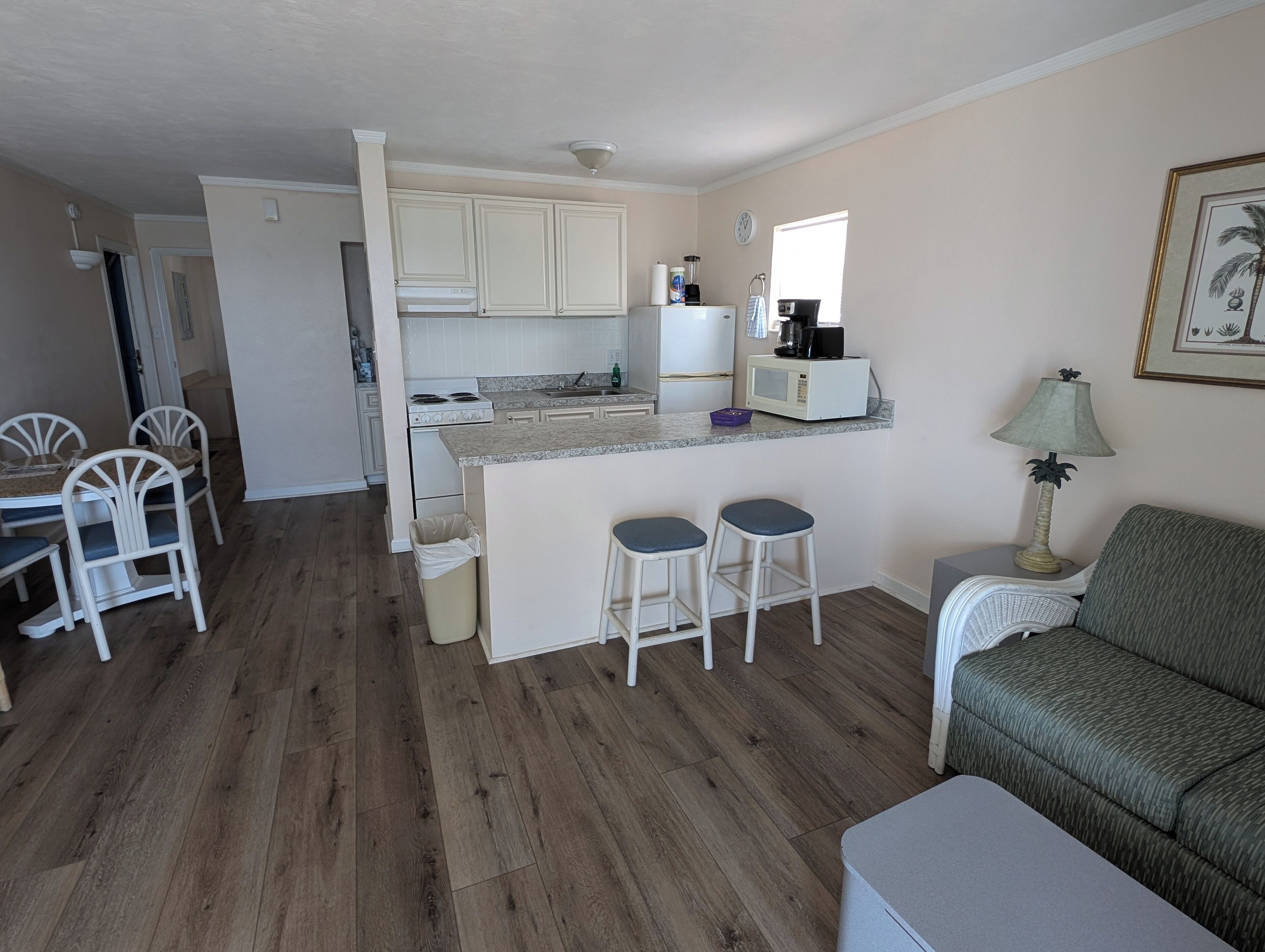 A compact kitchen with white cabinets, a microwave, and a coffee maker. There is a dining area with a table and chairs, and a green sofa in the living area.