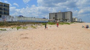 A beach with a large building in the background, a fence, and a few people. - Traders Inn Beach Club (Ormond Beach)