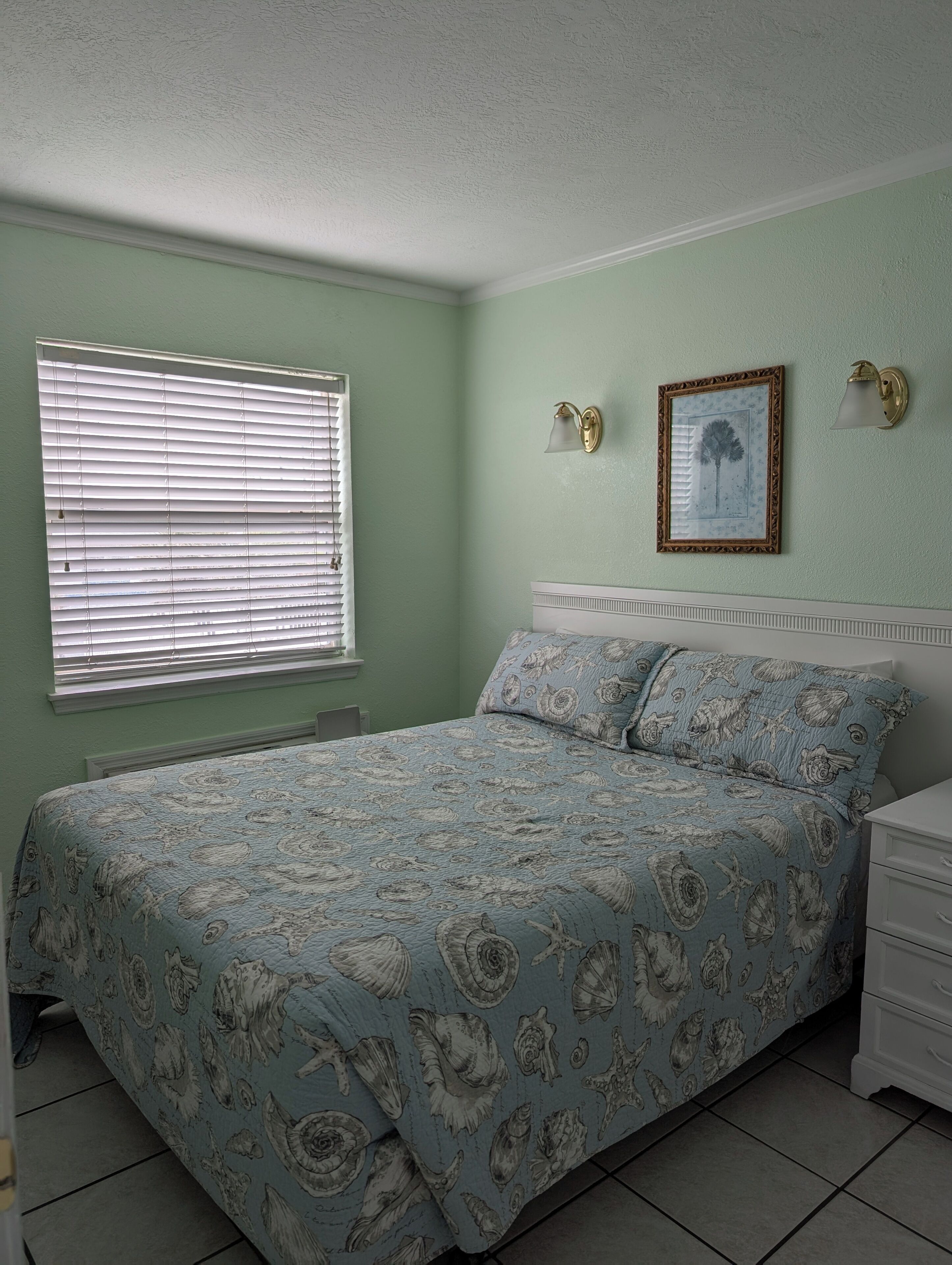 A bedroom with a bed, a nightstand, a wall-mounted lamp, and a framed picture.