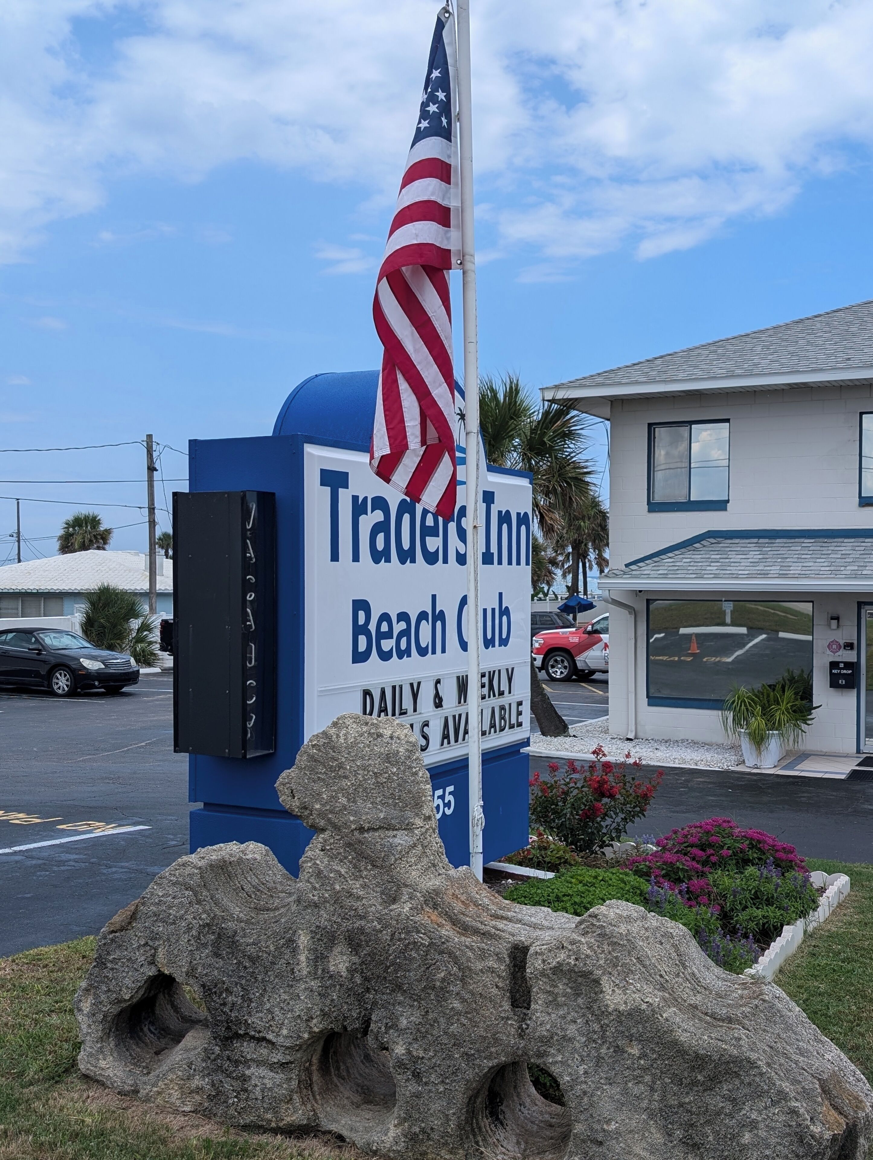 Une enseigne pour le Traders Inn Beach Club, avec un mât, une sculpture en pierre et un bâtiment en arrière-plan.