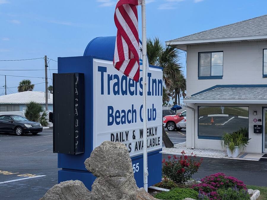 Un letrero del Traders Inn Beach Club, con un asta de bandera, una escultura de piedra y un edificio al fondo.