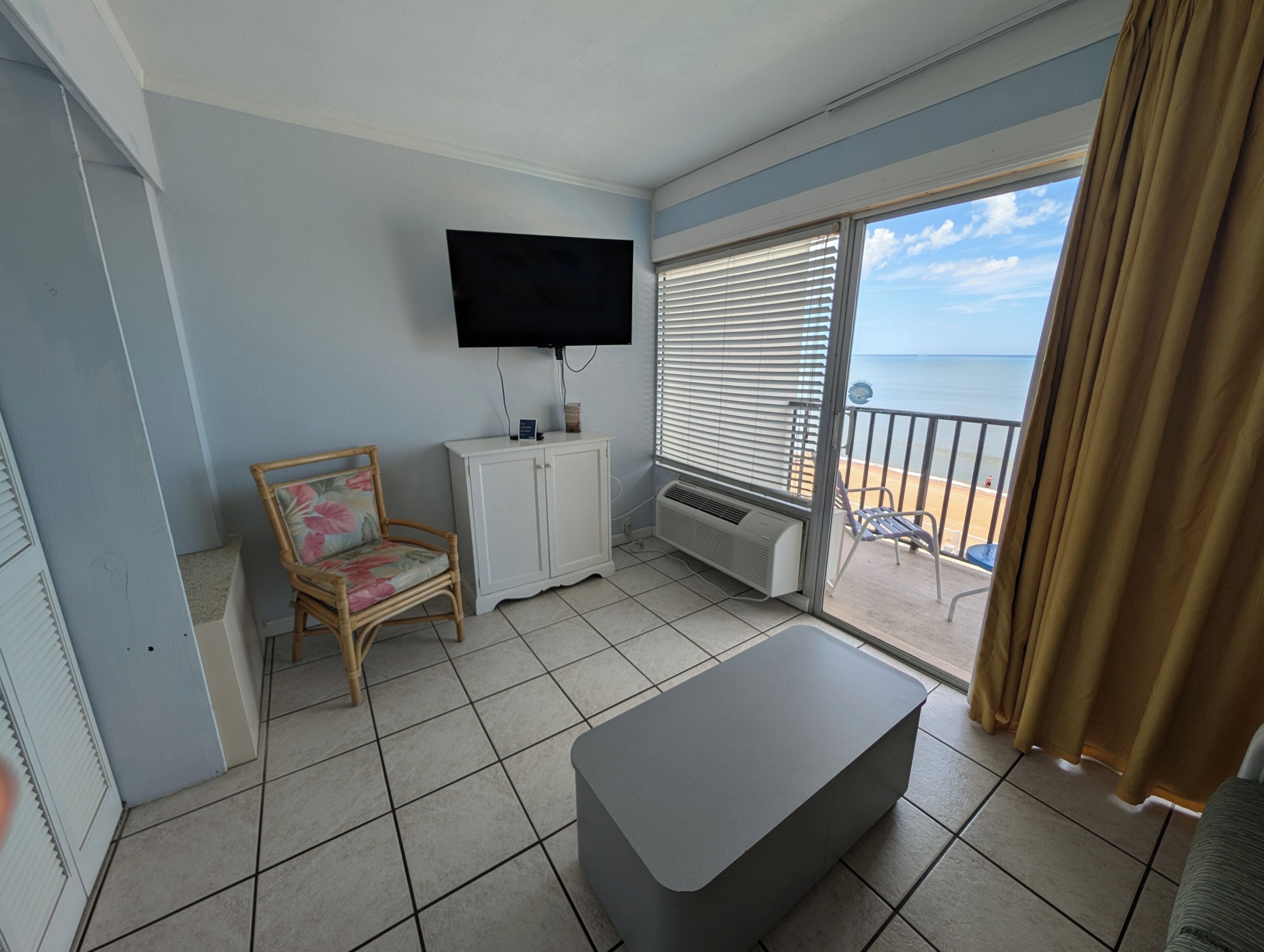 A balcony with a view of the sea, a flat-screen TV mounted on the wall, a white cabinet, a wicker chair, and a grey ottoman.
