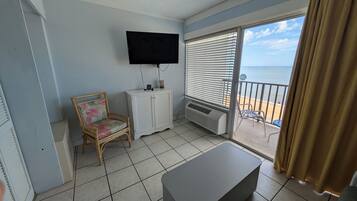 Un balcón con vistas al mar, un televisor de pantalla plana en la pared, un armario blanco, una silla de mimbre y un reposapiés gris.