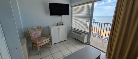 A balcony with a view of the sea, a flat-screen TV mounted on the wall, a white cabinet, a wicker chair, and a grey ottoman.