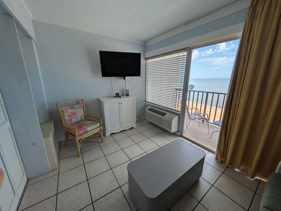 Un balcón con vistas al mar, un televisor de pantalla plana en la pared, un armario blanco, una silla de mimbre y un reposapiés gris.