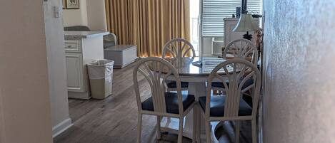 A dining area with a round table and white chairs, a kitchen in the background, and a window with blinds.