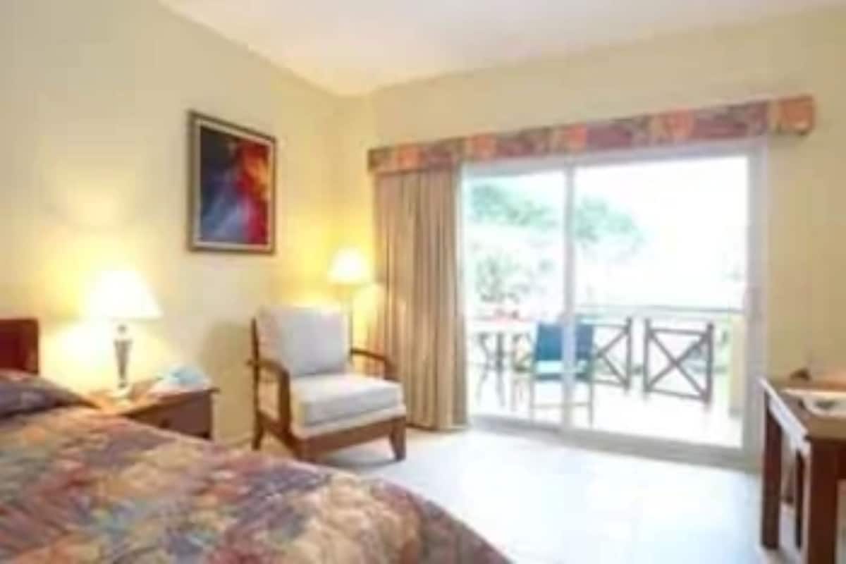 Een hotelkamer met een bed, een stoel, een tafel en een balkon met uitzicht op de omgeving.