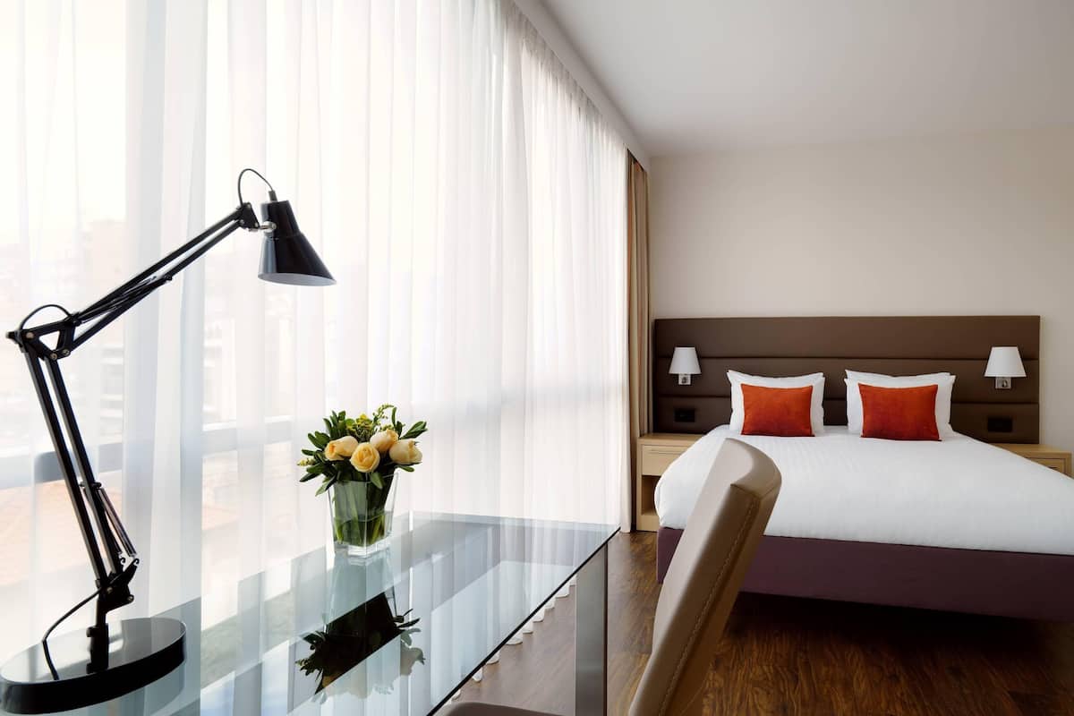 A hotel room with a bed, bedside tables, a desk with a lamp, and a vase with flowers.