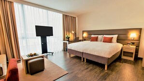 A modern hotel room with a large bed, a desk with a flat-screen TV, a seating area with a red chair, and a large window with curtains.