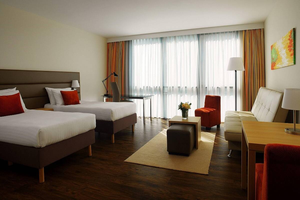 A hotel room with two beds, a desk, a chair, a sofa, and a lamp.