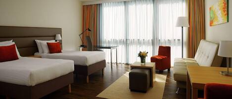 Quarto de hotel com duas camas, uma escrivaninha, uma cadeira, um sofá e um abajur.