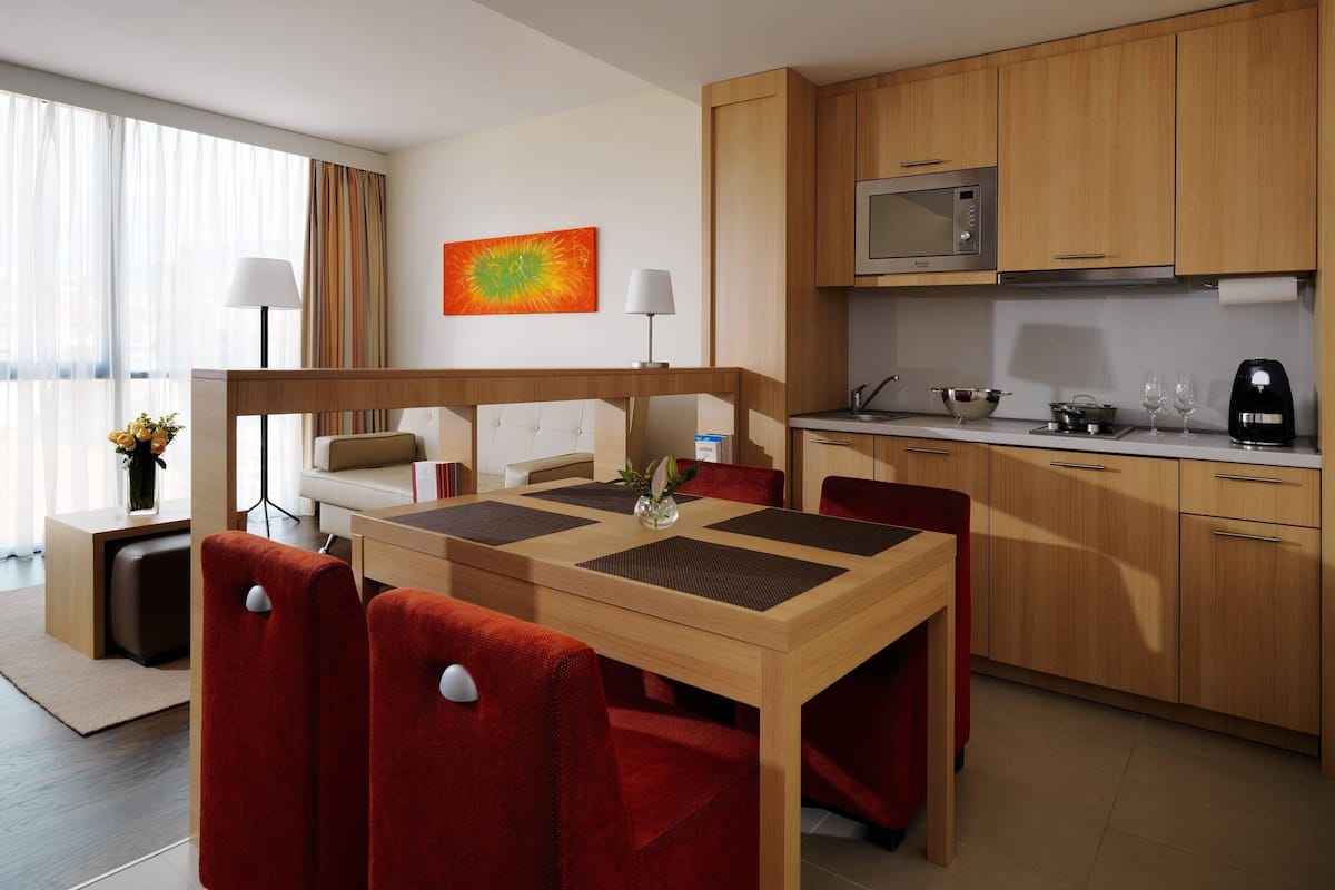 A modern hotel room with a dining area, a kitchenette, and a large window with curtains.
