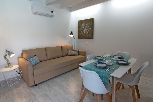 A modern living room with a sofa, dining table, and chairs. There is a lamp, a wall-mounted air conditioner, and a decorative compass wall art.