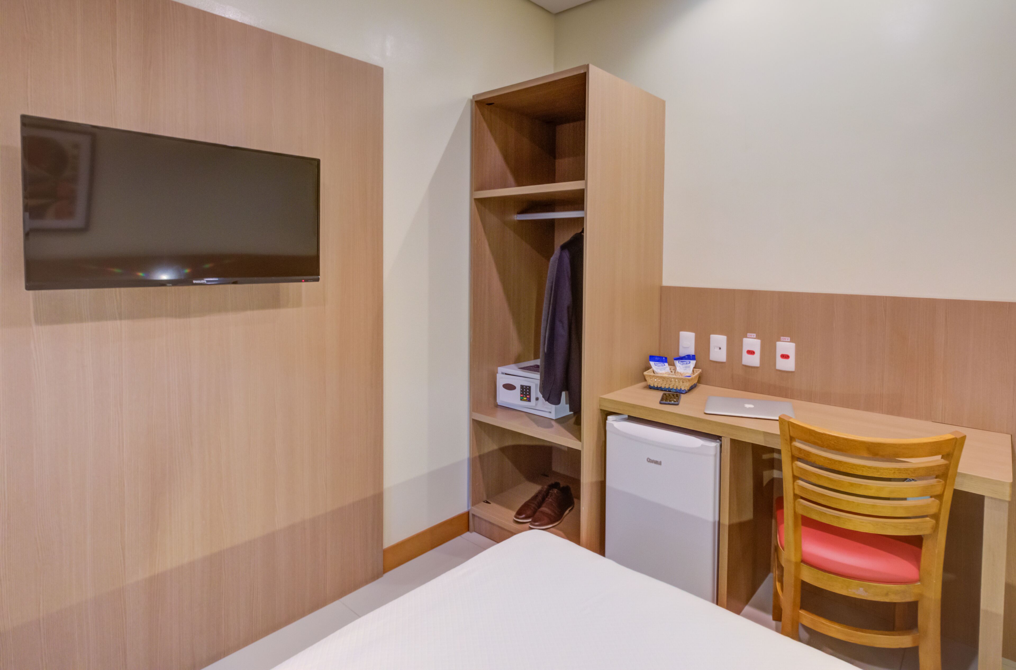 A compact hotel room with a bed, a desk with a computer, a small fridge, a wooden wardrobe, and a chair.