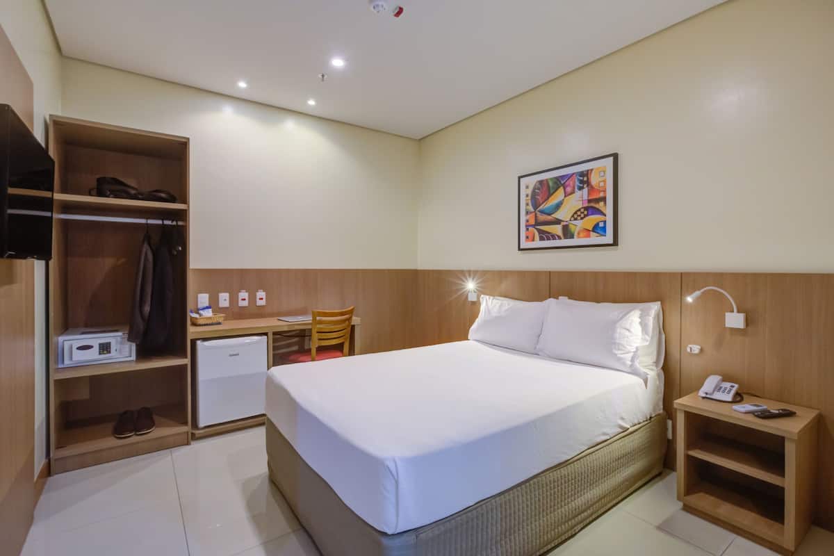 Une chambre d’hôtel comprenant un lit, un bureau avec une chaise, une armoire, un petit réfrigérateur et une table de chevet avec un téléphone.