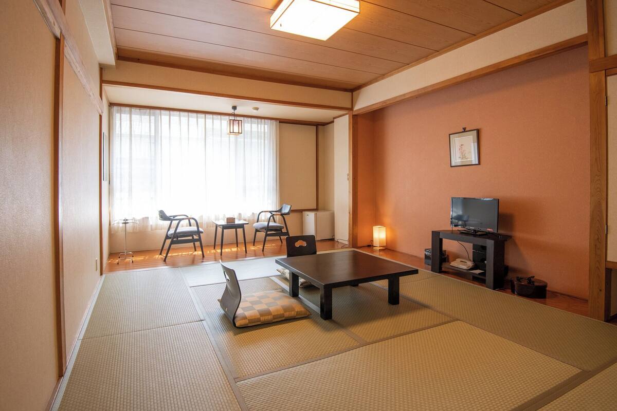 A traditional Japanese room with tatami flooring, a low table, chairs, a television, and a small cabinet.