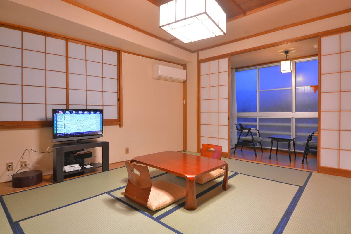 A traditional Japanese room with tatami flooring, a television on a low stand, and a sliding door leading to a balcony.