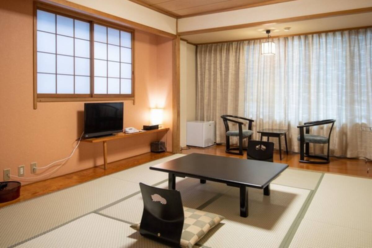 A traditional Japanese-style room with tatami flooring, a television, a desk, and a dining area with chairs and a table.