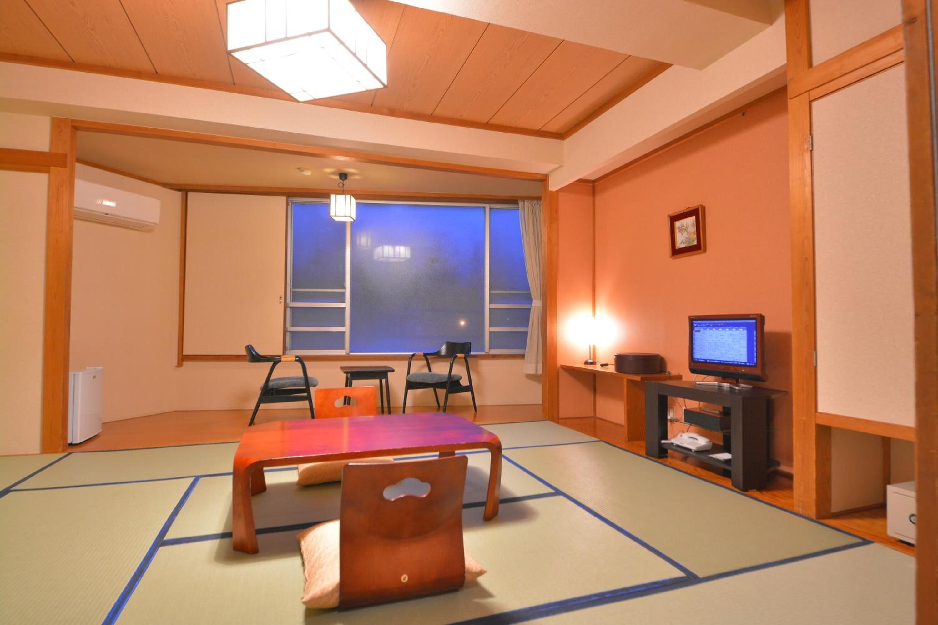 A traditional Japanese room with tatami flooring, a low table, chairs, a television, and a window with a view.