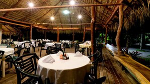 Un área de comedor al aire libre con mesas y sillas redondas, techo de paja y terraza de madera.