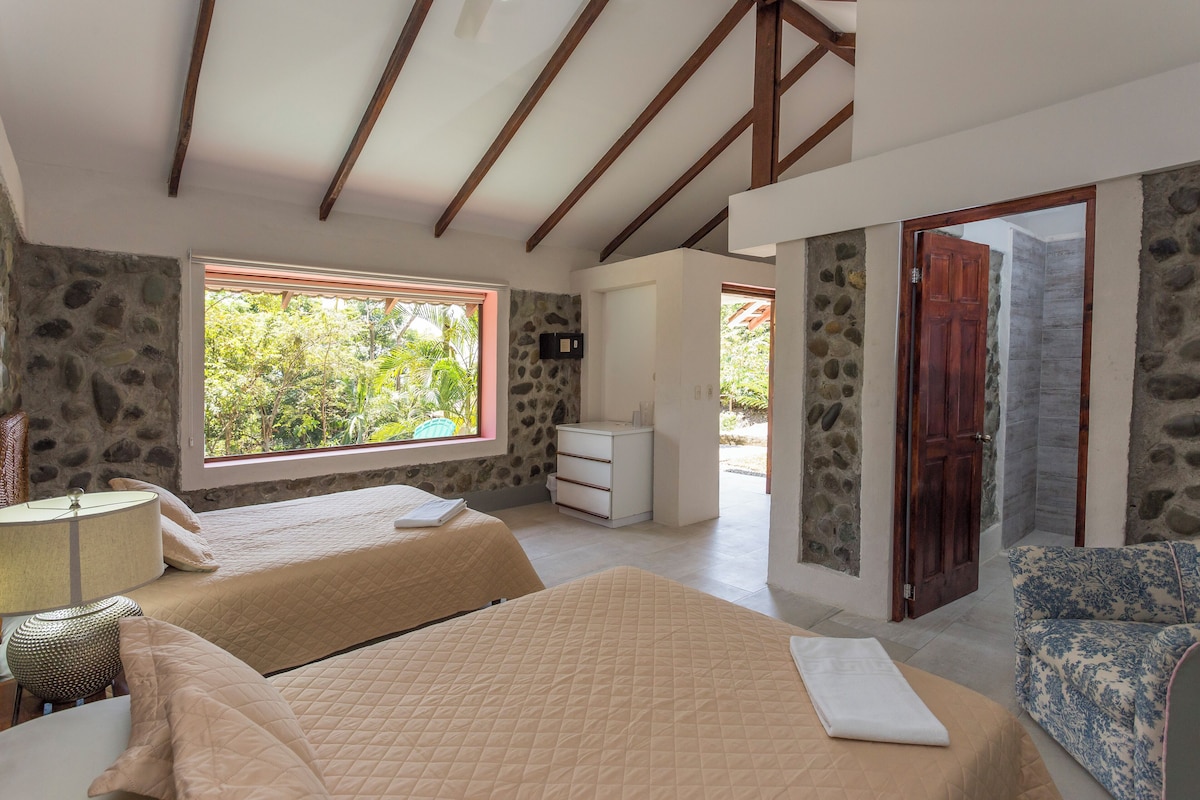 Un dormitorio con dos camas, una pared de piedra, techo de madera y ventana con vistas a la vegetación.