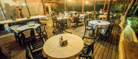 An outdoor dining area with round tables and chairs, a thatched roof, and a wooden deck.